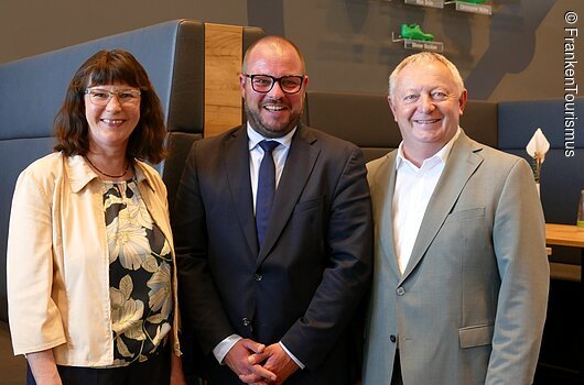 Geschäftsführerin Angelika Schäffer, Vorsitzender Sebastian Straubel und ehemaliger Vorsitzender Thomas Bold (v.l.n.r.) Drei Personen in Businesskleidung stehen nebeneinander und lächeln in einem modernen Raum mit Sitzbank und Tisch.