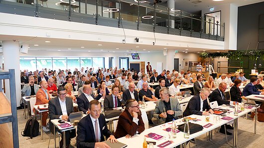 Großer Konferenzraum mit vielen sitzenden Personen bei einer Versammlung mit Tischen und Unterlagen.