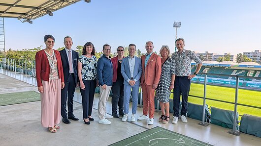 Team der Geschäftsstelle des Tourismusverbandes Franken e.V. Neun Personen stehen nebeneinander auf einer Stadiontribüne mit Blick auf ein Fußballfeld.