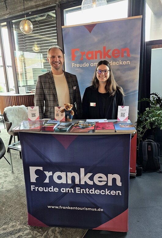 BayTM MICE Workshop 2026, Nürnberg Zwei Personen stehen hinter einem Infostand mit Broschüren und einem Plakat „Franken Freude am Entdecken“.