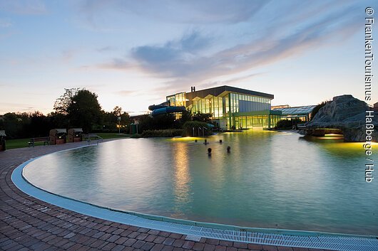 1. Naturheilwassersee Deutschlands in der FrankenTherme Bad Königshofen (Haßberge) Außenpool mit beleuchtetem Gebäude und Wasserrutsche bei Dämmerung, Menschen im Wasser sichtbar.