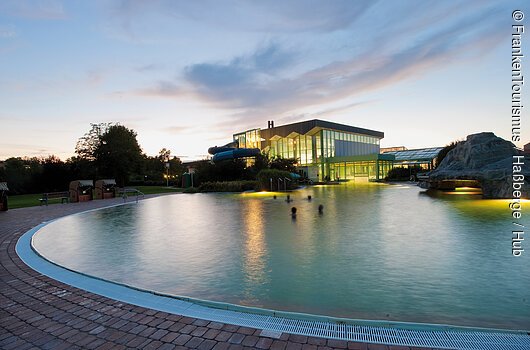 1. Naturheilwassersee Deutschlands in der FrankenTherme Bad Königshofen (Haßberge) Außenpool mit beleuchtetem Gebäude und Wasserrutsche bei Dämmerung, Menschen im Wasser sichtbar.