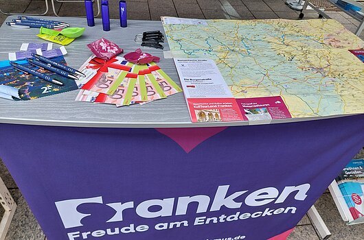 Auslage bei der FrankenTour 2026 Infostand mit Werbematerialien, Landkarte und Kugelschreibern auf Tisch mit lila Frankentuch