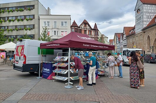 FrankenTour 2026, Reutlingen Menschen an einem Infostand mit Broschüren unter einem Zelt in einer Altstadt mit Fachwerkhäusern.