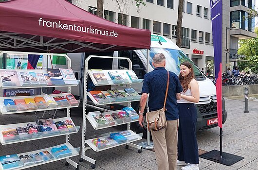 Infostand mit Prospekten unter rotem Zelt mit Aufschrift frankentourismus.de, zwei Personen im Gespräch davor.