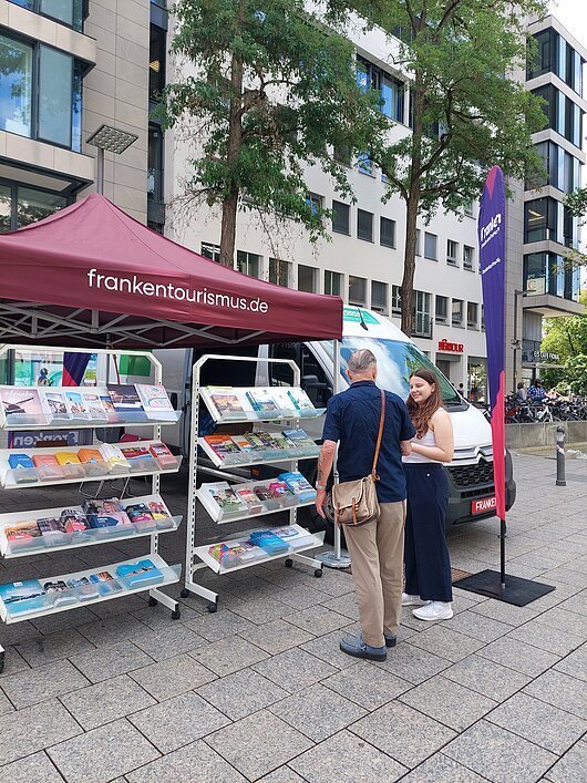 FrankenTour 2026, Ulm Infostand mit Prospekten unter rotem Zelt mit Aufschrift frankentourismus.de, zwei Personen im Gespräch davor.