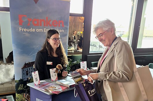 Zwei Frauen an einem Informationsstand mit „Franken Freude am Entdecken“-Werbung und Broschüren in einem Innenraum.