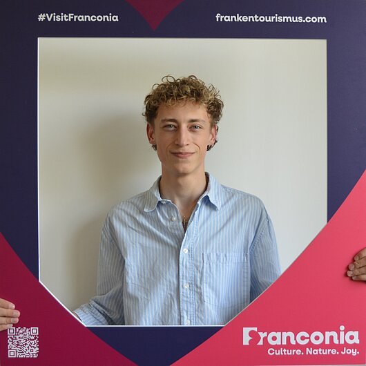 David Haller, Team FrankenTourismus Junger Mann mit lockigem Haar und blau-weiß gestreiftem Hemd hält einen Rahmen mit Aufschrift „Franconia“ und Webadresse.
