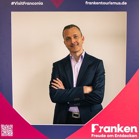 Alexander Hart, Team FrankenTourismus Mann in dunklem Anzug mit verschränkten Armen vor Rahmen mit Text #VisitFranconia und frankentourismus.de