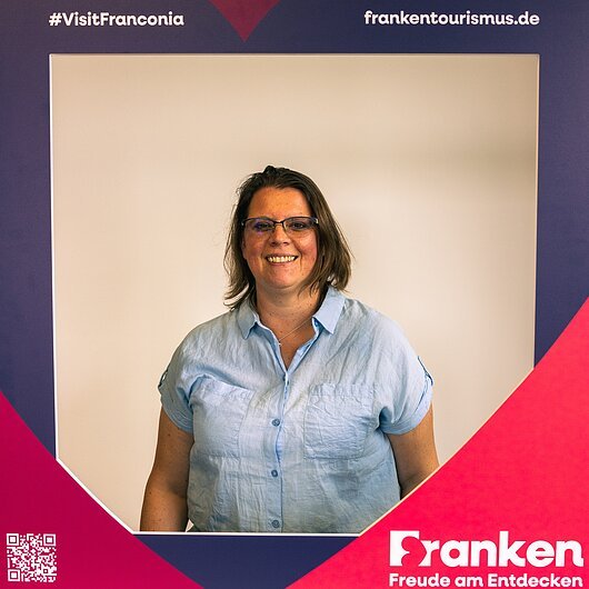 Claudia Büttner, Team FrankenTourismus Frau mit Brille und blauem Hemd lächelt vor Rahmen mit Text #VisitFranconia und frankentourismus.de
