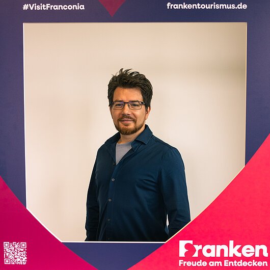 Manuel Teich, Team FrankenTourismus Porträt eines Mannes mit Brille und dunklem Hemd vor einem Rahmen mit Texten zu Franken-Tourismus und Hashtag VisitFranconia.