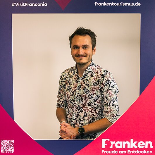 Lukas Miller, Team FrankenTourismus Porträt eines lächelnden Mannes mit gemustertem Hemd, umrahmt von einem Werberahmen mit Text zu Franken-Tourismus.